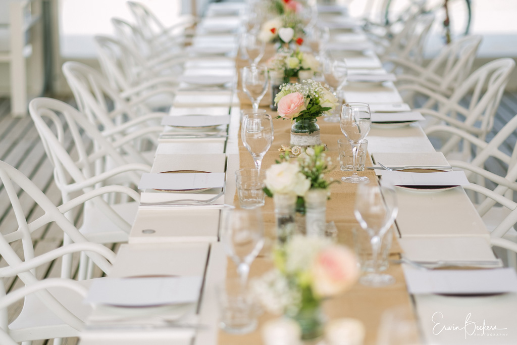 Lange, elegant gedekte bruiloftstafel met witte stoelen, bloemstukken en linnen loper bij Beachclub LaParade.